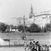 6. La chavalería se amontona alrededor del palo intentando lograr su premio. Como testigos, la Giralda y el antiguo edificio, hoy desaparecido, de la clínica del doctor Zbikowski. Década de 1960. ©ICAS-SAHP, Fototeca Municipal, archivo Cubiles.