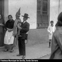 Hermandad de San Bernardo. Un padre lleva en brazos a su hijo vestido de nazareno en la avenida Menéndez y Pelayo. El niño porta un pequeño cirio y lleva el capirote levantado.  1925 La foto publicada ilustra un artículo El Noticiero Sevillano en que habla sobre la importancia de la continuidad en la tradición en el círculo familiar. 1925 ©ICAS-SAHP, Fototeca Municipal de Sevilla, fondo Serrano